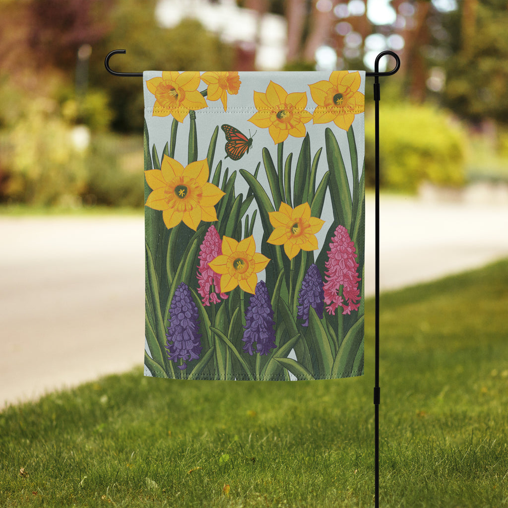 Hand-illustrated spring garden flag design with yellow daffodils, pink and purple hyacinths, green leaves, and a monarch butterfly on a soft blue background.