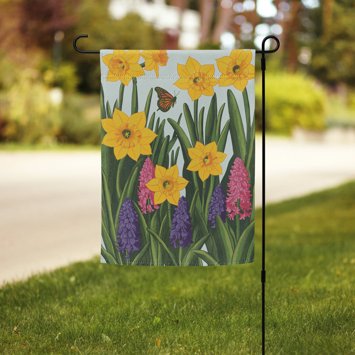 Hand-illustrated spring garden flag design with yellow daffodils, pink and purple hyacinths, green leaves, and a monarch butterfly on a soft blue background.